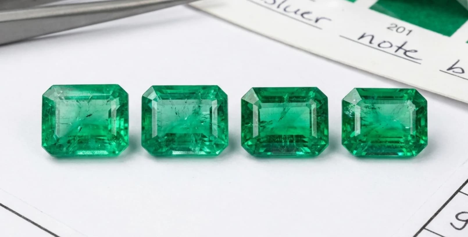 Four emerald-cut emeralds laid out in a row on white grading paper for comparison.