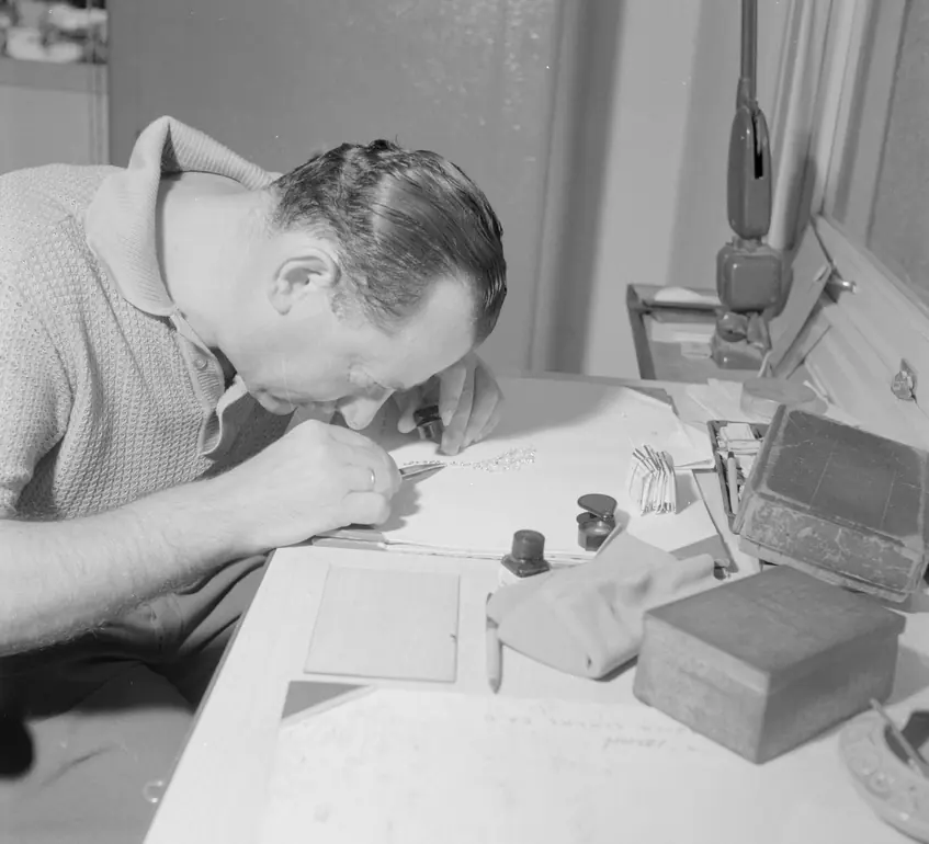 A man at a bench examining a small stone through a loupe, parcel papers spread in front of him.