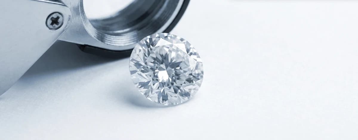 A loose round brilliant diamond resting beside an open jeweller’s loupe on a pale ground.