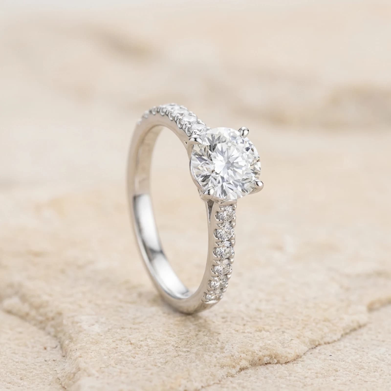 A white metal solitaire ring with a diamond-set band, standing upright on a pale stone ground.